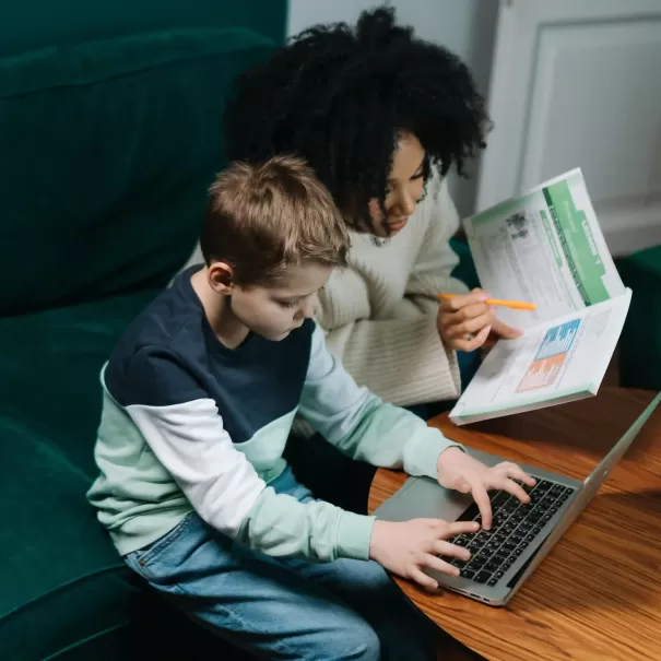 Teacher with pen and book sat on sofa with male child student who is on a laptop