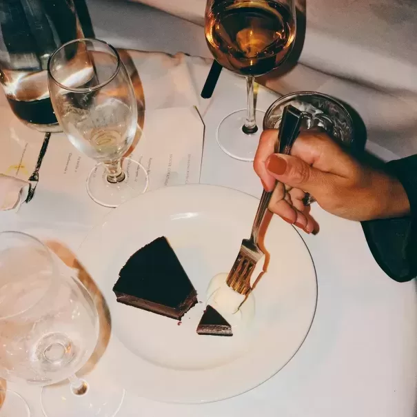 A hand holds a fork with a bite of chocolate cake and cream over a white plate. Two slices of chocolate cake, several empty glasses, and a glass of wine are on the white tablecloth.