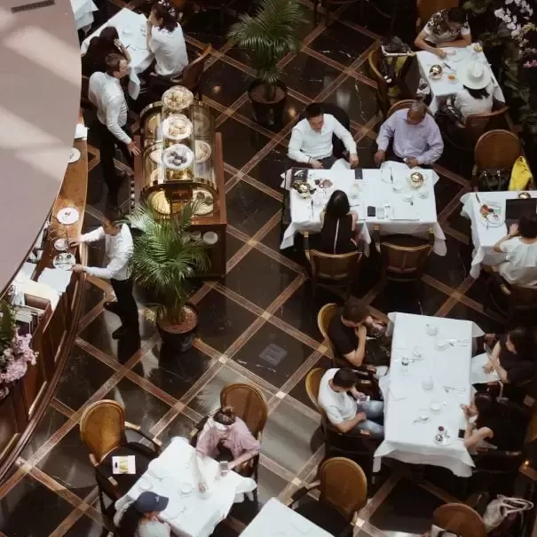 Birdseye view of people sat at rectangular dining tables with white table clothes in a restaurant with waitors and brown chequered floors