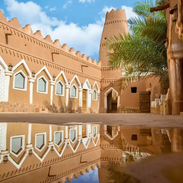 A traditional mud-brick building with white-trimmed windows and a tall tower is reflected in a puddle under a blue sky with clouds, alongside a palm tree in Saudi Arabia.