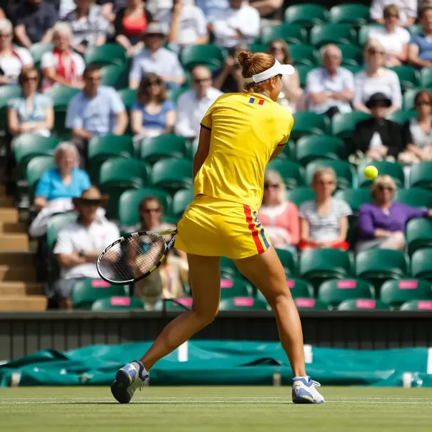 A tennis player in a yellow outfit with colorful stripes hits a backhand shot on a grass court during a match. Spectators in casual summer attire watch from the stands in the background.