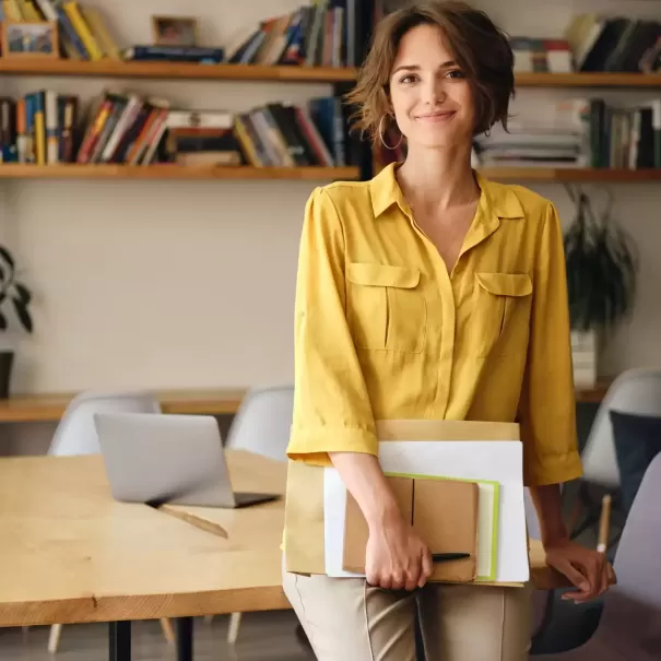 Women in classroom in yellow blouse sat with paperwork and notebook smiling on a desk