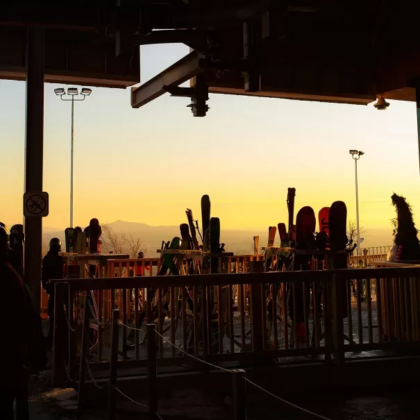 Ski racks holding various skis and snowboards stand under a lift station at sunset, with people nearby and distant mountains silhouetted against an orange sky.