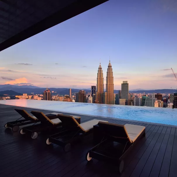 Four empty lounge chairs face an infinity pool overlooking a city skyline at sunset, with the Petronas Twin Towers prominently visible in the background.