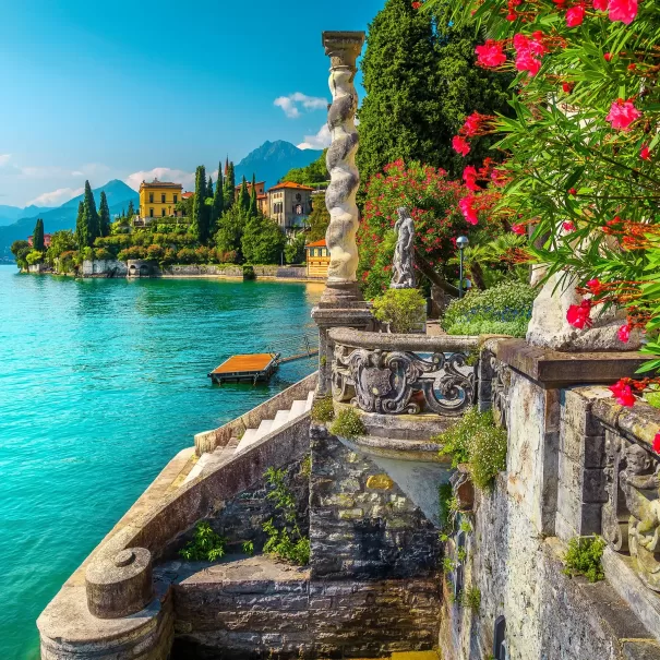 Scenic view of Lake Como with a stone balustrade and vibrant flowers in the foreground. Elegant villas are nestled among lush greenery along the waters edge, with mountains in the background under a clear blue sky.