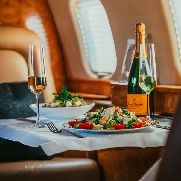 A gourmet meal with salad, a glass of champagne, and a bottle of Veuve Clicquot is served on a white tablecloth inside a luxurious private jet with leather seats and sunlight streaming through the windows.
