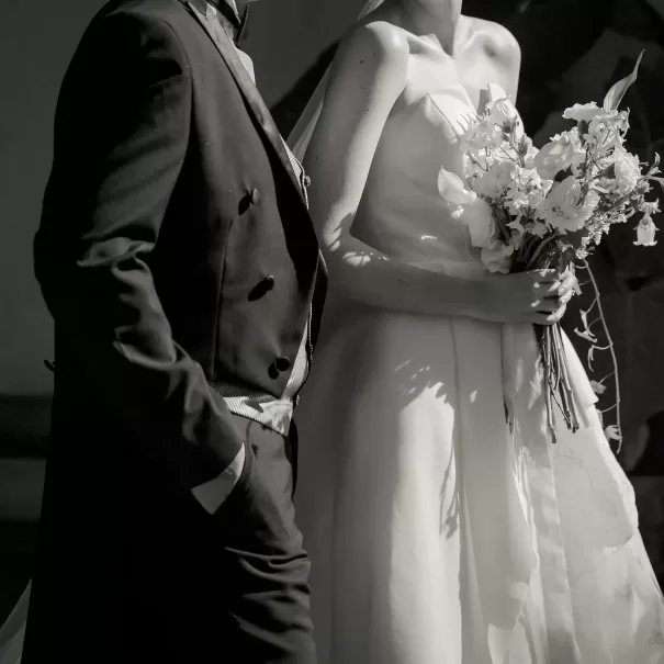 A black-and-white photo of a bride in a strapless wedding dress holding a bouquet of flowers, standing beside a person in a dark suit with one hand in their pocket. Their faces are not visible.
