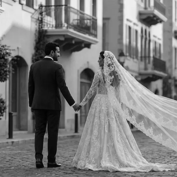 A bride and groom hold hands while walking down a cobblestone street. The bride wears a long, lace wedding gown with a veil. The groom is in a suit. The scene is in black and white, with old buildings in the background.