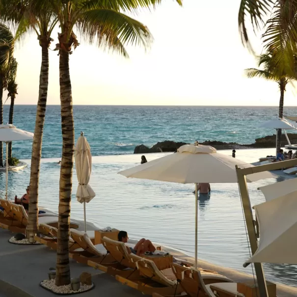 People relax on lounge chairs by an infinity pool next to the ocean, shaded by palm trees and white umbrellas, with a few swimmers in the pool and waves in the background at sunset.