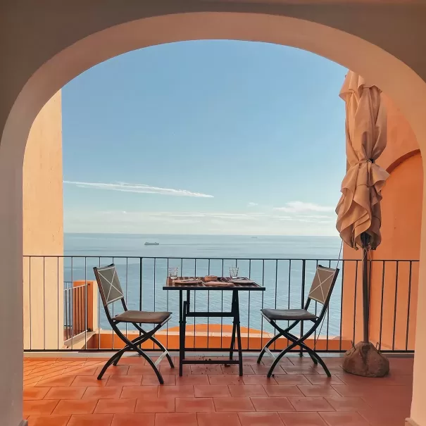 A small table set for two sits on a terrace with orange tiles, overlooking a calm blue sea. Two empty chairs face the ocean, framed by an arched doorway. A closed umbrella stands by the railing under a clear sky.