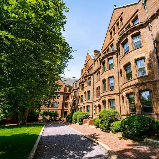 A path lined with trees and bushes leads to historic brown brick apartment buildings with many windows, bathed in sunlight on a clear day.