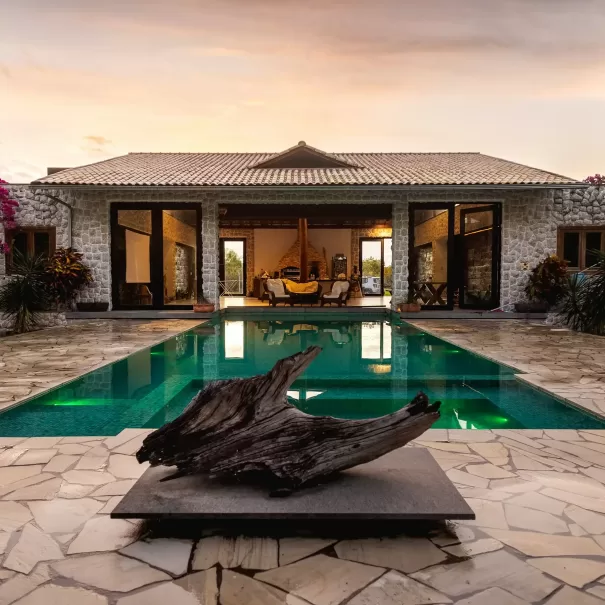 A luxurious villa with stone walls and large glass doors opens onto a tranquil swimming pool, reflecting the warm sunset sky. A sculptural piece of driftwood sits on a stone platform at the pool’s edge.