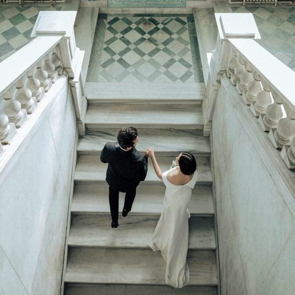 A bride in a white dress and a groom in a dark suit hold hands as they walk up marble stairs in an elegant building, seen from above.