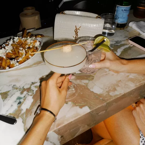 Two women seated at a marble table toast with cocktails; one holds a martini with an olive, the other a drink with lime. A plate of fries with cheese and a white YSL bag are visible on the table.