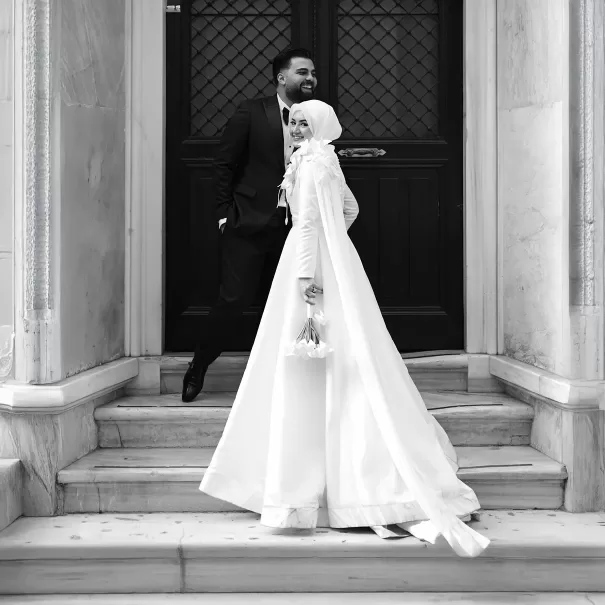 A smiling bride in a white hijab and gown holds a bouquet while standing on marble steps, with a groom in a suit standing behind her, both in front of ornate double doors. Black and white photo.