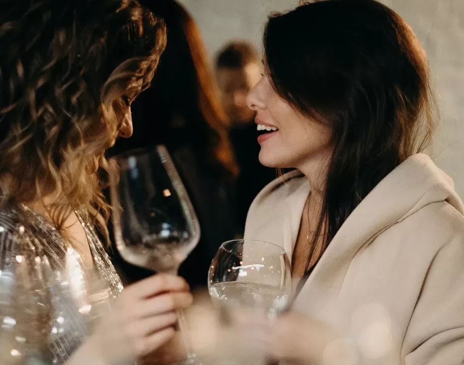 Two women at a social gathering hold empty wine glasses and smile at each other while talking. The background is softly blurred, highlighting their interaction.