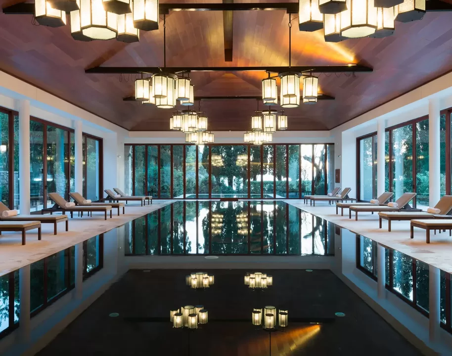 Luxurious indoor pool with lounge chairs along both sides, large windows letting in natural light, and modern hanging lanterns reflecting in the still water. Warm ambient lighting and wooden ceiling create a serene, inviting atmosphere.