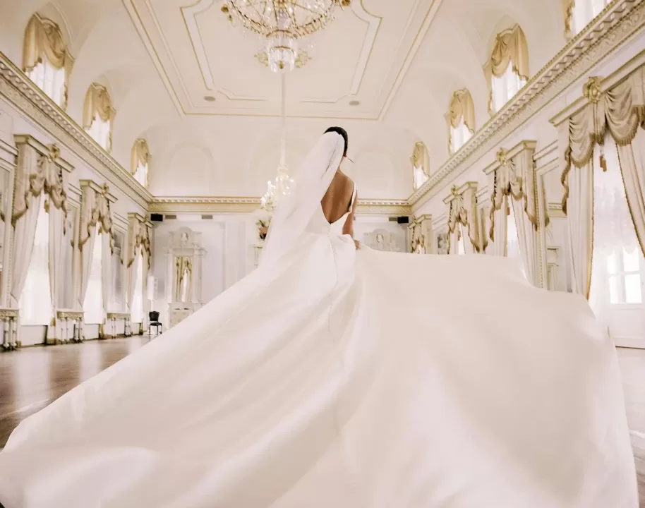 Dramatic shot of the back of a bridge wearing long trailed white, silky wedding gown and vail in a white ballroom with gold chandelier, trims and curtains.