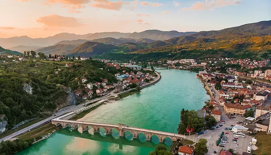 Aerial view of a scenic town by a winding turquoise river, with a historic stone bridge and lush green mountains in the background under a sunset sky. The town features clusters of houses and buildings along the riverbanks.