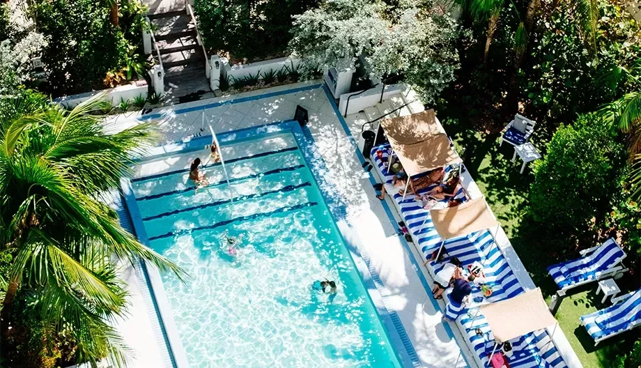 Aerial view of a vibrant swimming pool surrounded by lush greenery. People are swimming and lounging on blue and white striped chairs under tan umbrellas. The scene conveys a relaxing, tropical atmosphere.
