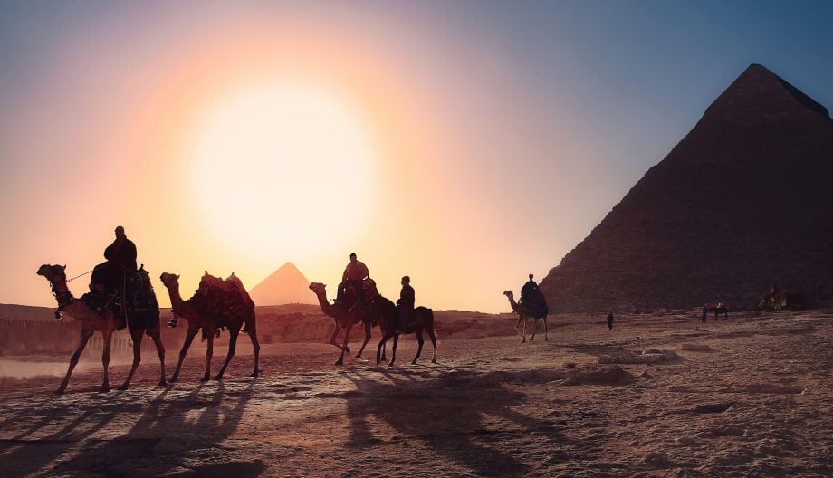 Silhouetted against a setting sun, a caravan of camels with riders travels across a desert, with the Great Pyramids of Giza in the background. The sky is a gradient of warm colors, creating a dramatic and picturesque scene.