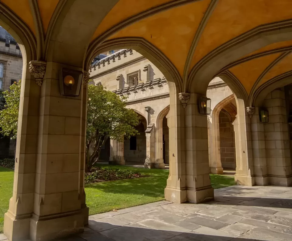 A historic stone archway leads to a courtyard with green grass and a tree. The structure features a vaulted ceiling and ornate detailing. Sunlight creates shadows on the ground, enhancing the architectural elegance of the University of Melbourne, Australia.