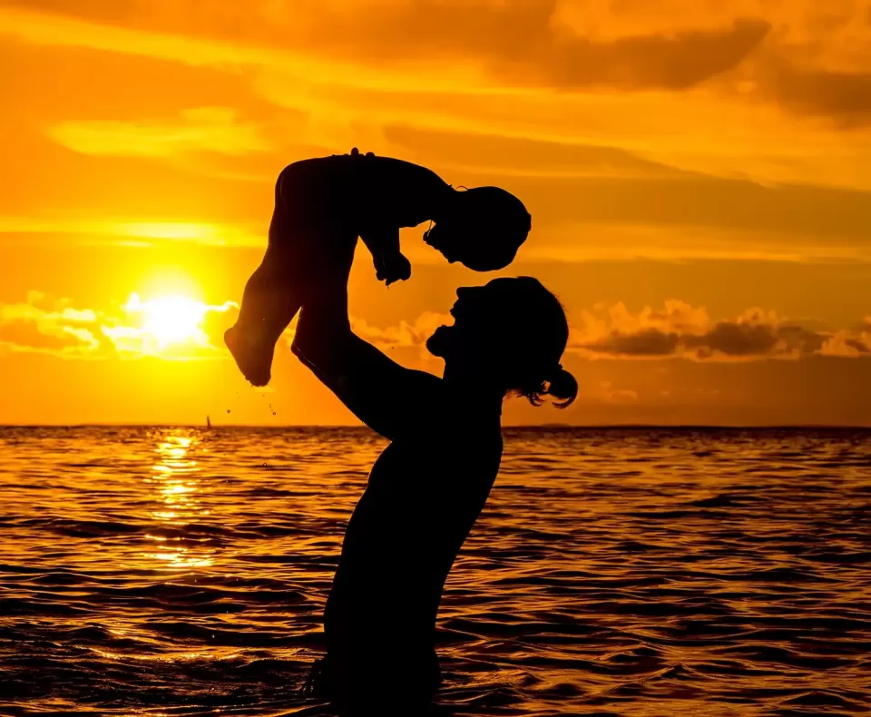 Silhouette of a person lifting a child against a vibrant sunset over the ocean. The sky is a mix of orange and yellow hues, with the sun near the horizon. The scene conveys warmth and joy, highlighting the bond between the two figures.