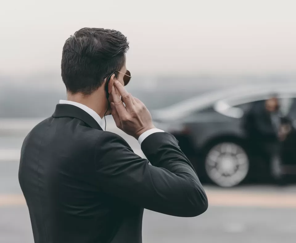 Man in suit with hand up to headpiece in ear looking at a luxury car