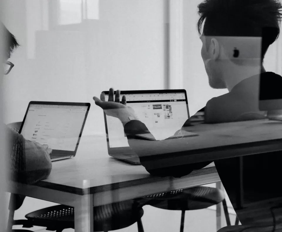 Two people sat at a desk on laptops talking