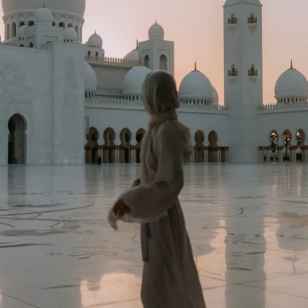 A person in modest clothing stands on a reflective marble courtyard at sunset, near the white domes and minarets of a grand mosque, likely the Sheikh Zayed Grand Mosque.