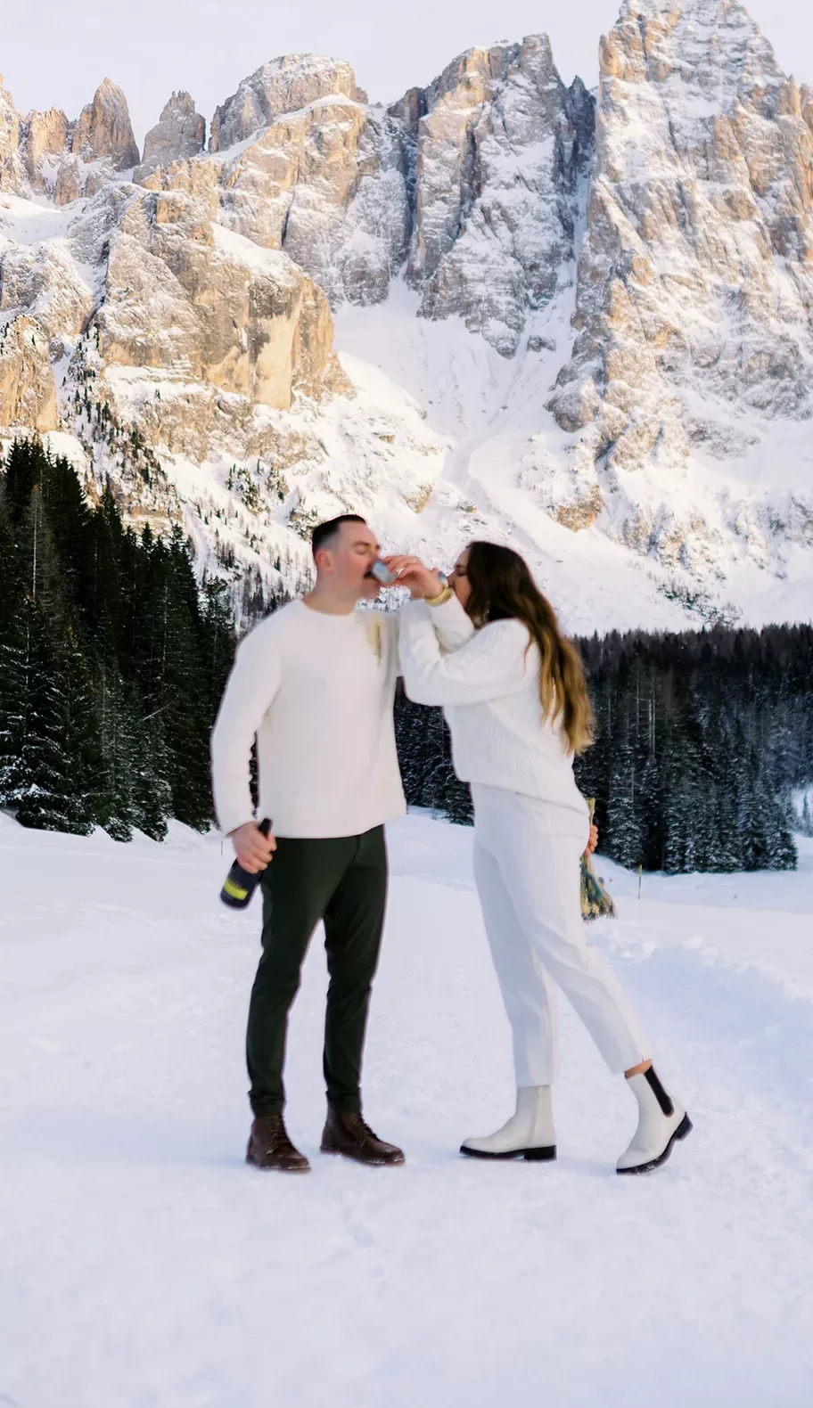 A couple dressed in white and dark pants stands on snowy ground in front of tall, snow-covered mountains and pine trees, sharing a drink and smiling. The man holds a bottle while the woman playfully drinks from a glass.