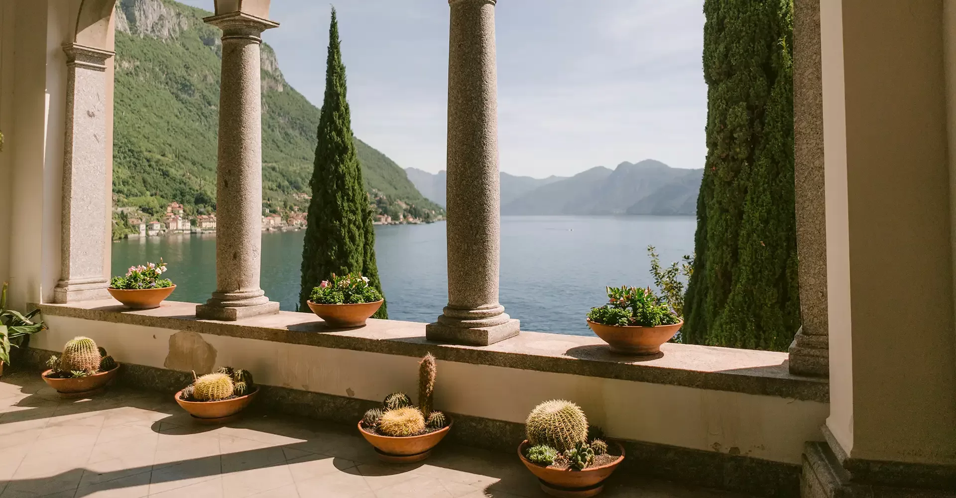 Stone columns frame a scenic view of a lake and distant mountains, with potted plants and cacti arranged along the terrace ledge. Tall cypress trees stand outside, adding to the tranquil, Mediterranean setting.