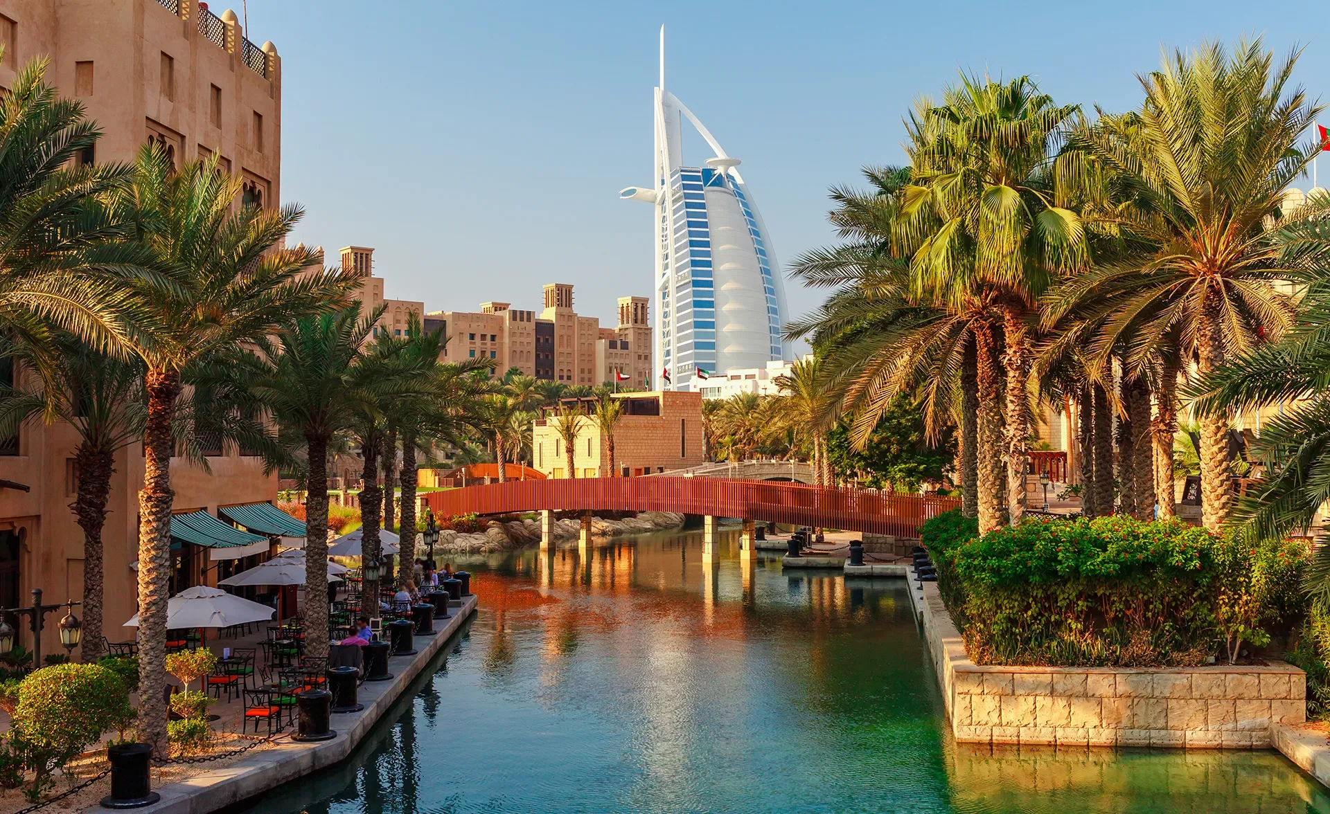 A canal lined with palm trees and outdoor cafes, with a wooden bridge crossing over the water and the sail-shaped Burj Al Arab hotel visible in the background under a clear blue sky in Dubai.