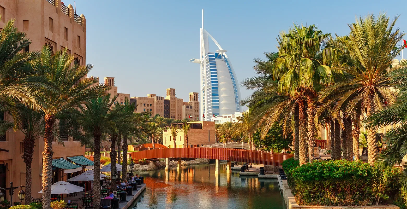 A canal lined with palm trees and outdoor cafes, with a wooden bridge crossing the water, and the sail-shaped Burj Al Arab hotel in the background under a clear blue sky.