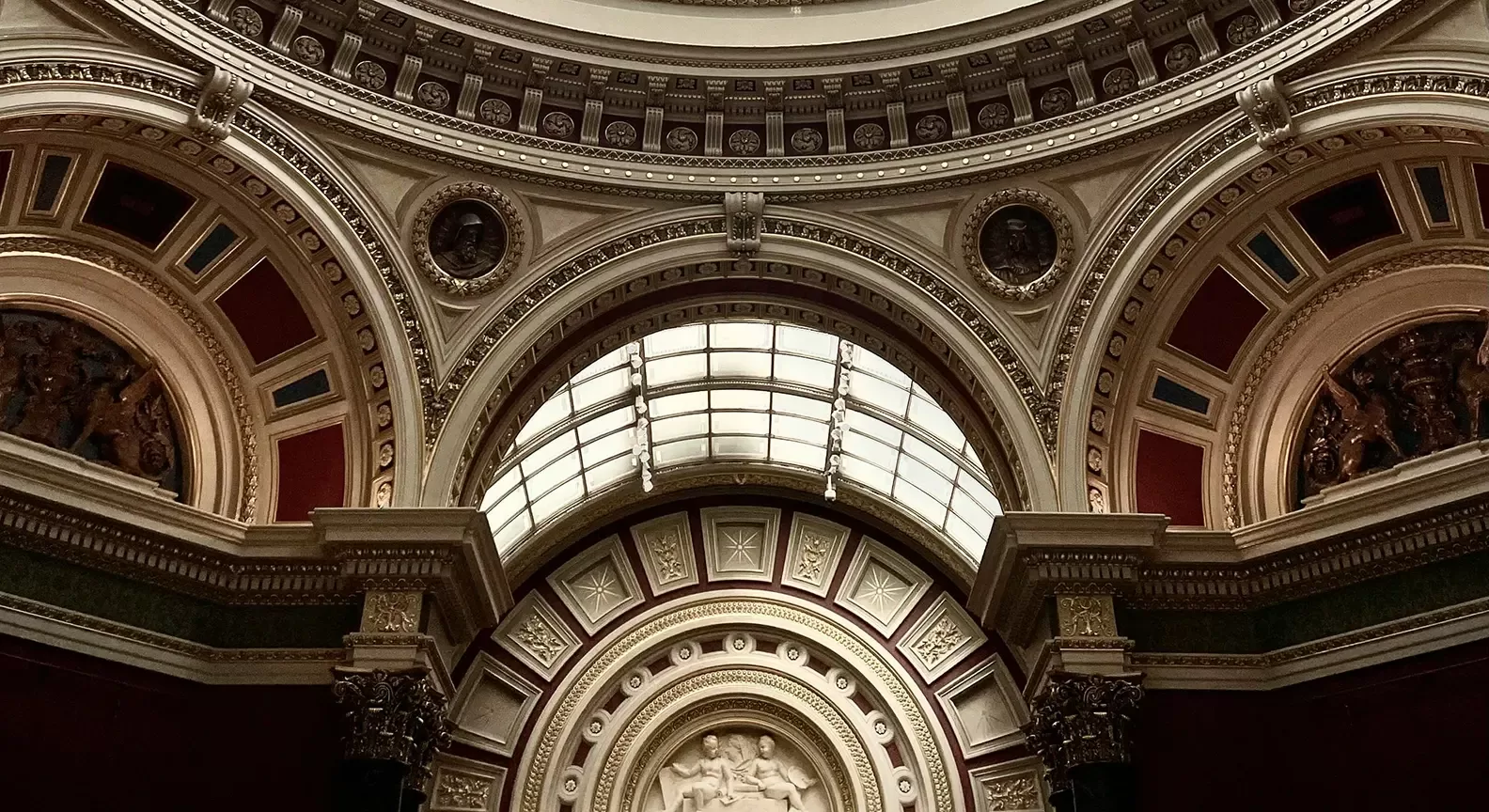 An ornate domed ceiling with arches, classical columns, detailed carvings, and sculpted reliefs. The ceiling features a large skylight and decorative circular medallions in a richly adorned architectural interior.
