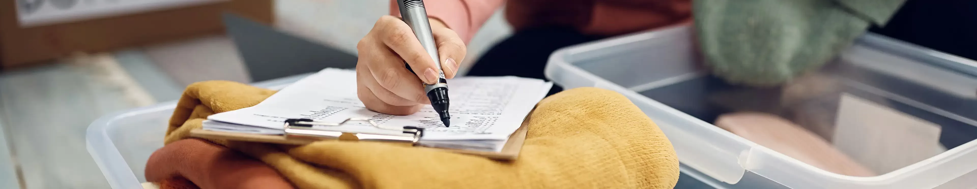 A person holding a pen writes on a clipboard with papers, positioned over a transparent storage container filled with folded clothes.