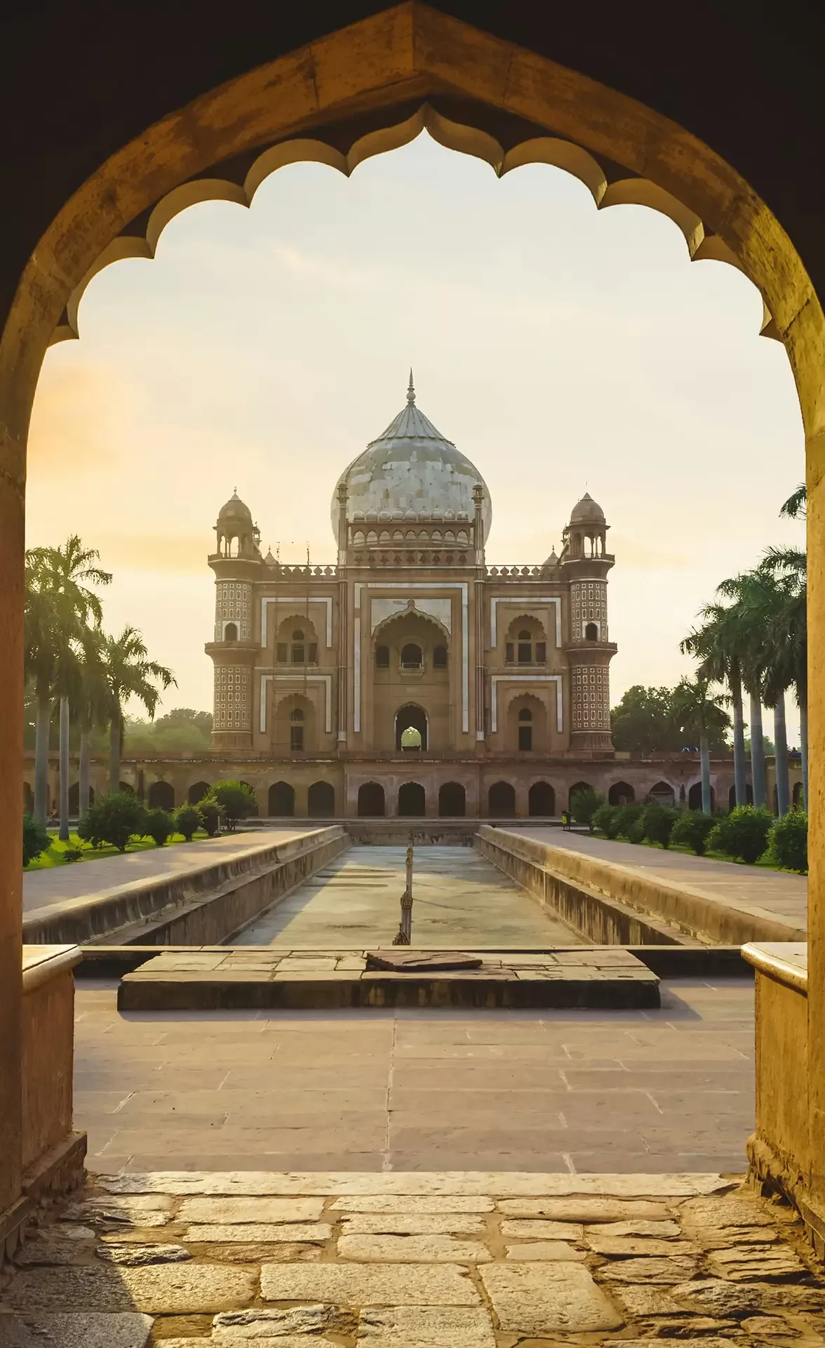 A view of Humayun’s Tomb in Delhi, India, framed by an arched entrance with a pathway and water channel leading toward the historic Mughal structure at sunrise.