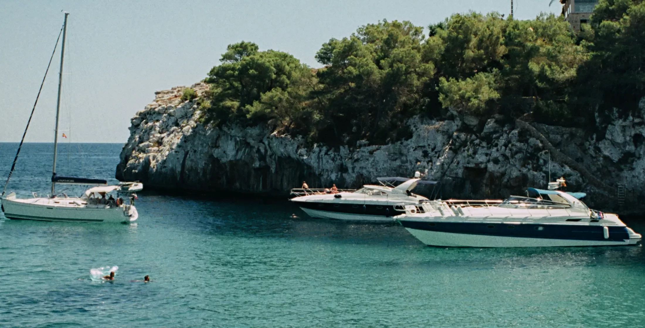 Two boats are anchored in clear turquoise water near a rocky coastline with green trees. A swimmer is visible in the water, and a sailboat floats nearby under a sunny sky.