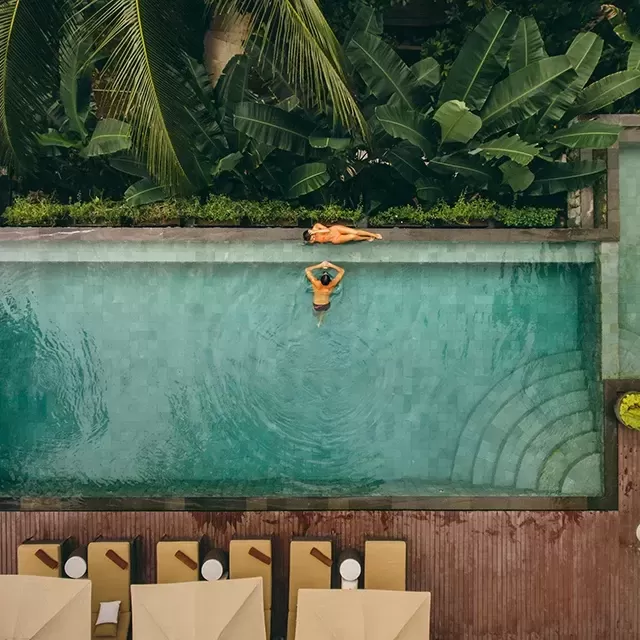 Birdseye view of outdoor swimming pool and loungers, amongst huge green forest trees with a women bathing on the edge of the pool and a man inside of the pool.