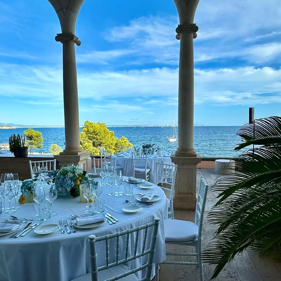 Elegant outdoor dining setup on a terrace overlooking the ocean, with round tables, white chairs, blue floral centerpieces, and clear blue sky with scattered clouds in the background.