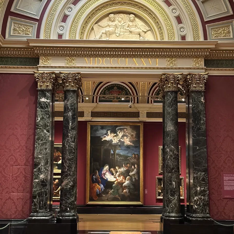 A grand museum hall with ornate gold decorations, red walls, marble columns, and a large classical painting displayed under an arched ceiling inscribed with MDCCCLXXVI. Smaller paintings are visible on the sides.