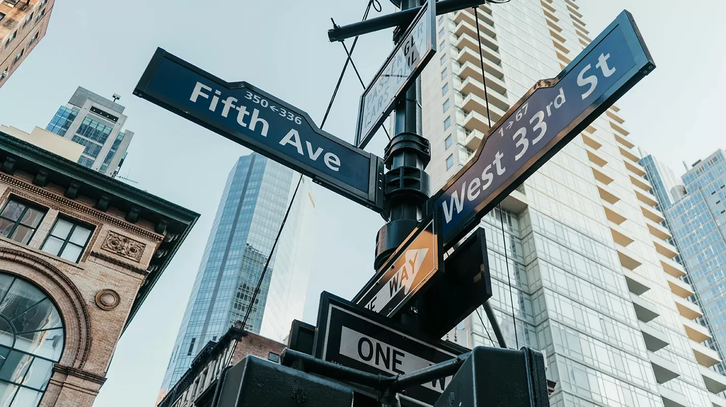 Street signs for Fifth Ave and West 33rd St mounted on a pole, surrounded by tall buildings and skyscrapers. The view is angled upwards, capturing a mix of historical and modern architecture against a clear sky.