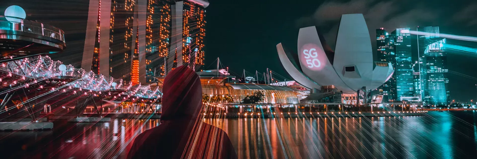 Person wearing a red hoodie looking across the river at the lit up city of Singapore