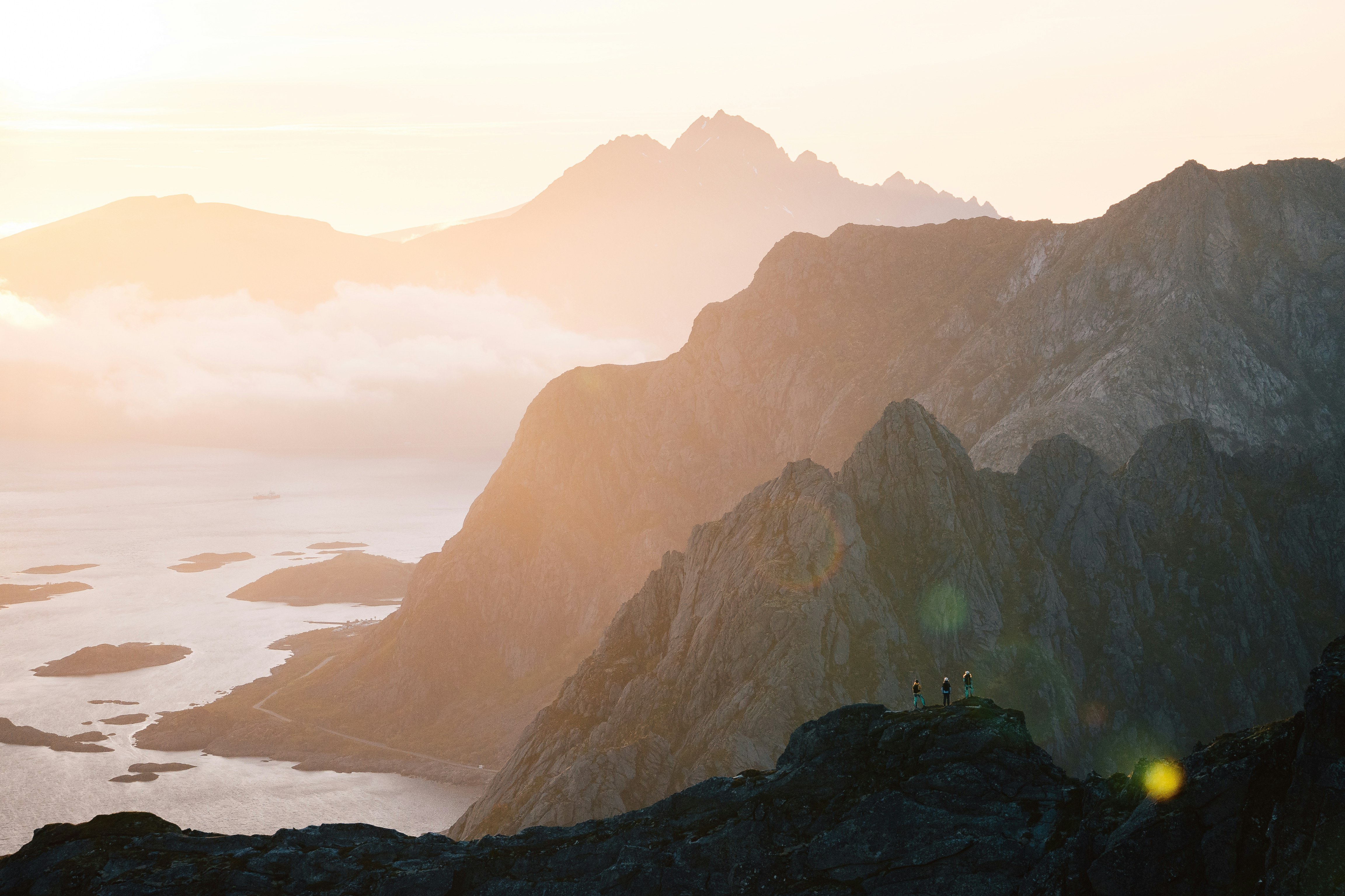 A golden sunrise illuminates rugged mountains and a serene, winding coastline. Three small hikers stand on a rocky peak in the foreground, admiring the expansive, misty landscape.