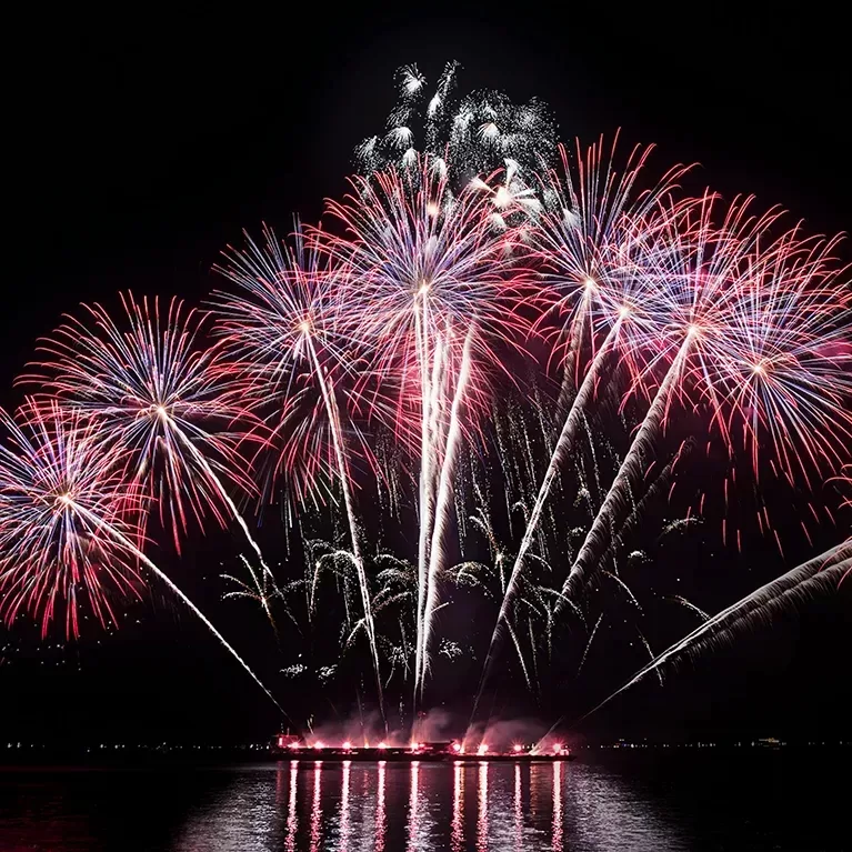 A vibrant fireworks display lights up the night sky over a body of water, with red, white, and blue pyrotechnics creating a stunning visual show. The reflections on the water add to the spectacle.