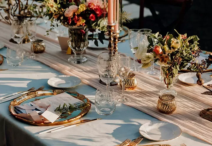 Elegant table setting with gold-rimmed plates, glasses, candles, and floral centerpieces on a pale tablecloth and fabric runner, creating a warm and inviting atmosphere for a formal meal.