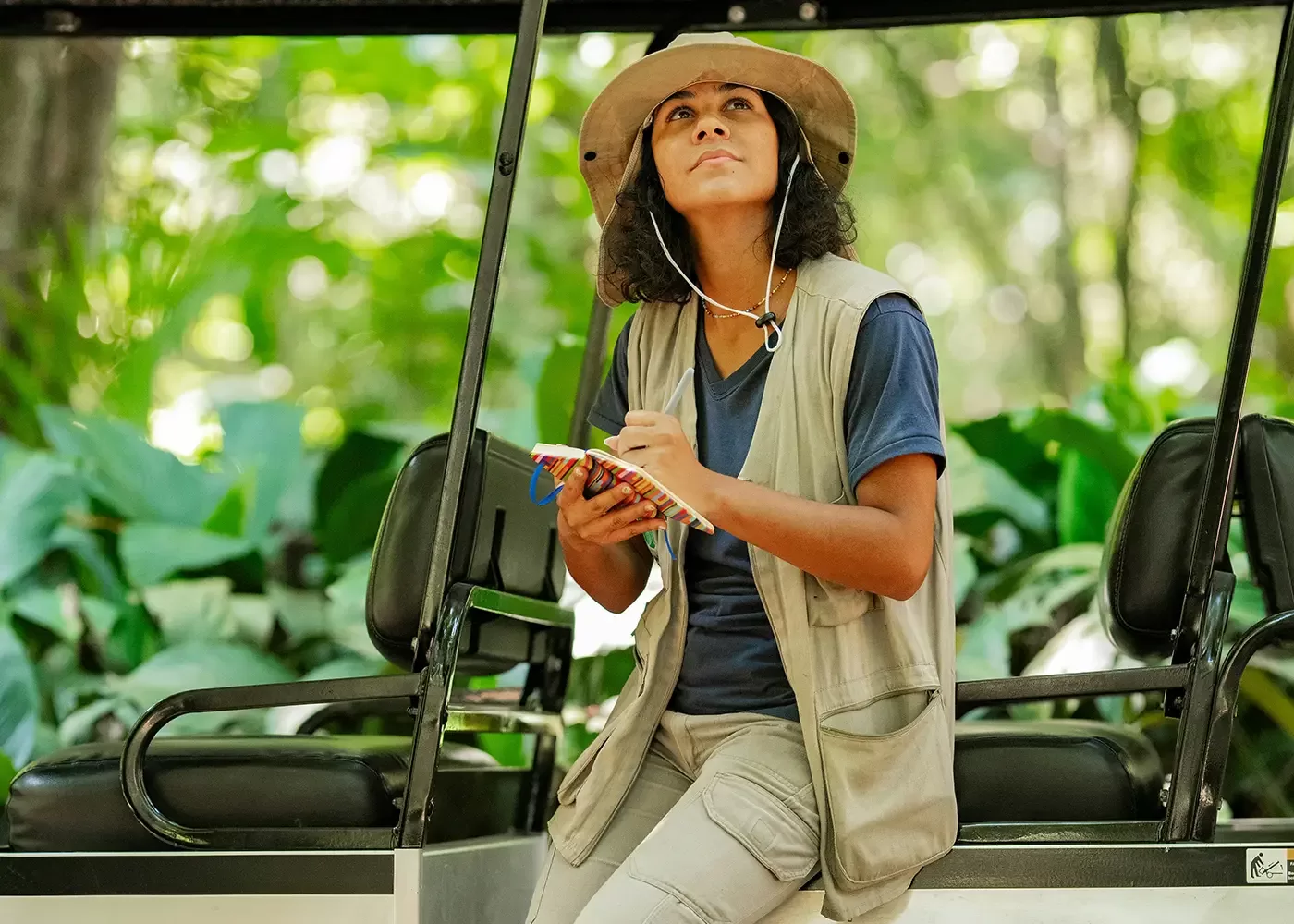 A person wearing a safari hat and vest sits in an open vehicle, holding a notepad and pen, looking up thoughtfully at lush green foliage in a forest or jungle setting.