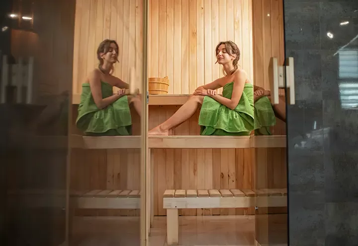 A woman wrapped in a green towel sits on a wooden bench inside a sauna, with her reflection visible on the glass door.