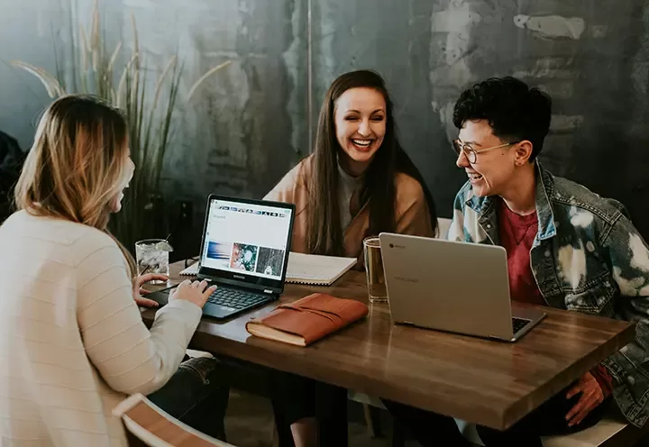 Three people sit at a table with laptops, notebooks, and drinks, smiling and laughing together in a relaxed, casual setting with a dark wall and a plant in the background.