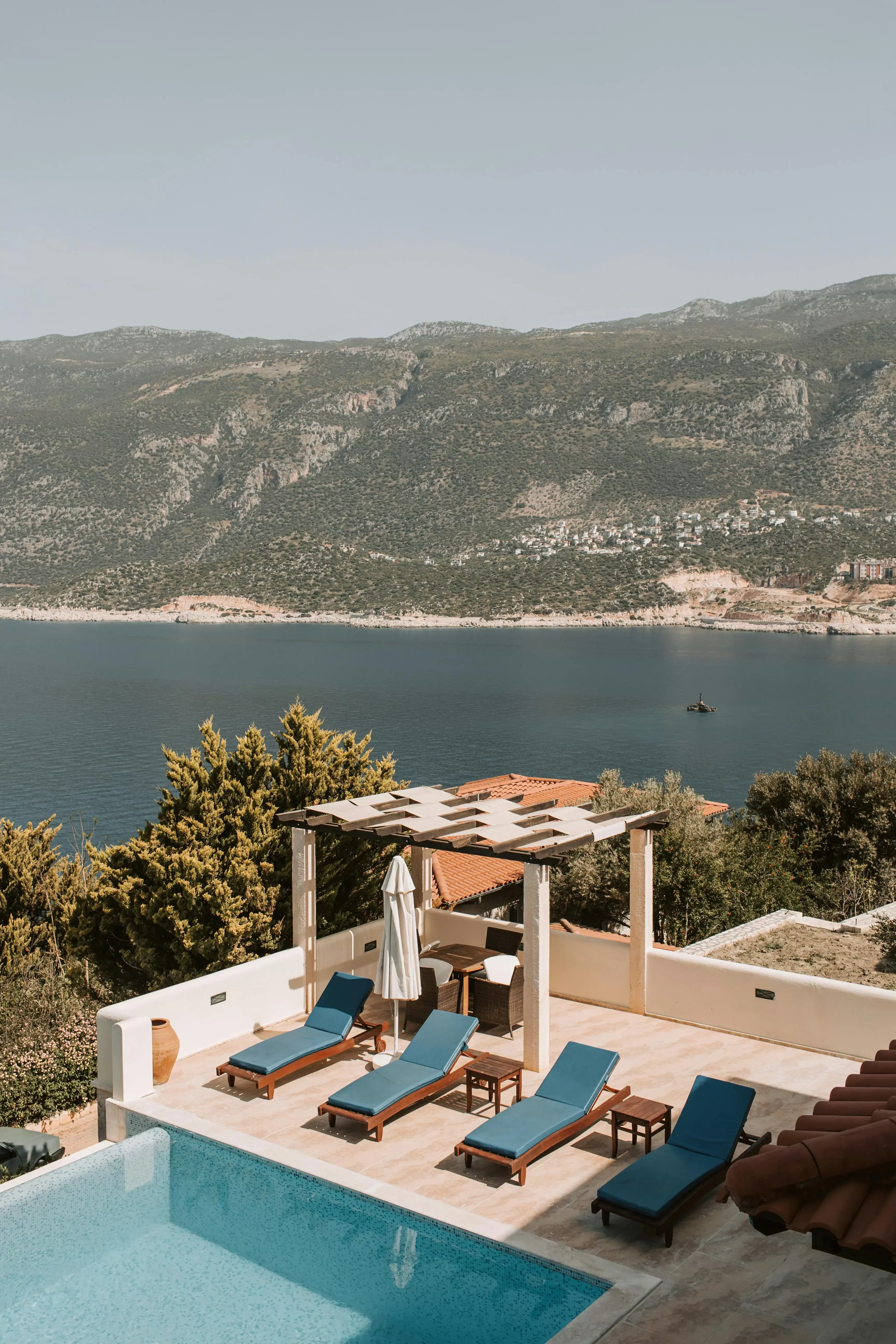 A poolside patio with four blue lounge chairs under a pergola overlooks a calm sea and distant green mountains on a sunny day.